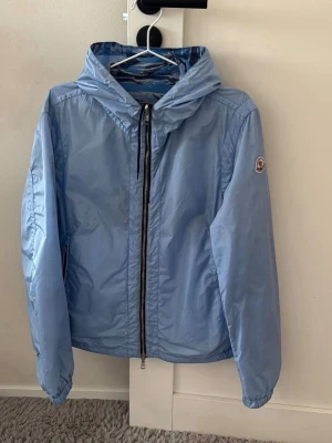 Moncler wind jacket - Snygg vändbar vindjacka från Moncler i ljusblått på ena sidan och blått kamouflagemönster på andra. Jackan har huva, dragkedja framtill och Moncler-logga på ärmen. Tillverkad i lätt, vindtätt material och har elastiska muddar vid ärmslut.