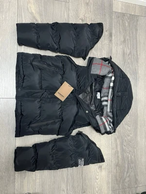 Svart dunväst från Burberry - Snygg svart dunväst från Burberry med avtagbara ärmar och huva. Insidan av huvan har klassiskt rutigt Burberry-mönster i grått, vitt och rött. Västen har dragkedja, tryckknappar och två fickor framtill. Perfekt för lager-på-lager och streetstyle. Passar S och M.