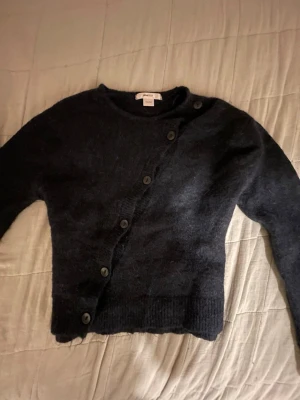 Svart kofta med sned knäppning - Mysig svart kofta med unik sned knäppning framtill och rund hals. Koftan har långa ärmar och ribbade muddar vid ärmslut och nederkant. Tillverkad i mjukt, fluffigt material som känns skönt mot huden. Perfekt för lager på lager och en cool detalj i garderoben.