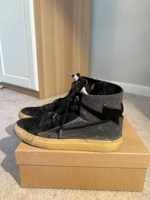 Zadig & Voltaire svarta high top sneakers - Snygga high top sneakers från Zadig & Voltaire i svart mocka och textil med glittriga detaljer och blixtformad patch på sidan. Skorna har svarta skosnören, dragkedja på insidan och beige sula för en cool kontrast.