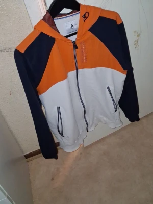 Färgglad Ship Master hoodie - Säljer en hoodie från Ship Master med dragkedja, huva och fickor. Färgerna är orange, marinblå och vit i blockmönster. På ryggen står det 'SHIP MASTER' i stora bokstäver. Perfekt för dig som gillar sportig och avslappnad stil.