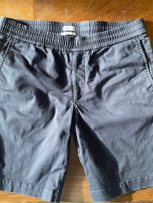 Svarta shorts från Filippa K - Snygga svarta shorts från Filippa K med elastisk midja och sidofickor. Klassisk rak passform och två bakfickor. Perfekta för sommardagar och enkla att matcha med olika stilar.