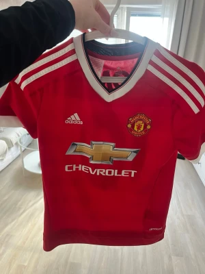 Manchester United matchtröja Adidas - Röd Manchester United matchtröja från Adidas med vita detaljer och tre ränder på axlarna. Tröjan har klubbmärke, Adidas-logga och Chevrolet som sponsortryck på bröstet. Tillverkad i funktionsmaterialet ClimaCool för bra ventilation.