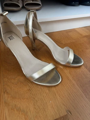 Guldiga pumps med öppen tå från XIT - Snygga guldiga pumps från XIT med öppen tå och smal rem över foten. Skorna har en elegant ankelrem och smal klack som ger en stilren look. Tillverkade i ett glansigt syntetmaterial som ger en lyxig känsla. Perfekta för att lyfta vilken outfit som helst.