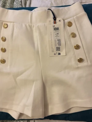 Vita shorts med guldknappar XS - Stilrena vita shorts från Lindex med hög midja och sex dekorativa guldknappar framtill. Klassisk och elegant design med raka ben och slät front. Perfekta för sommarens alla tillfällen.