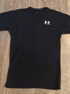 Svart t-shirt från Under Armour - Säljer en svart t-shirt från Under Armour med korta ärmar och logotyp på bröstet. Perfekt för dig som gillar sportiga och stilrena plagg. Materialet är syntet och t-shirten har en normal passform.