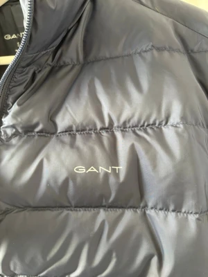 Svart pufferjacka från GANT - Snygg svart pufferjacka från GANT med diskret logga på bröstet. Jackan har hög krage, dragkedja framtill och quiltad design. Tillverkad i ett glansigt material som ger en modern look och håller dig varm under kalla dagar.