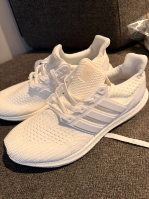 Vita Adidas Ultraboost sneakers - Snygga helvita Adidas Ultraboost sneakers med stickad ovandel och klassiska tre ränder på sidan. Skorna har en bekväm och flexibel sula samt vita skosnören. Perfekta för dig som gillar stilrena och sportiga sneakers.