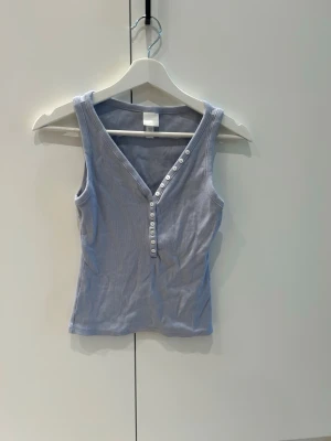 Ljusblå ribbad linnetopp H&M - Snygg ljusblå ribbad linnetopp från H&M med v-ringning och dekorativa knappar framtill. Toppen är ärmlös och har en smickrande passform som passar perfekt till jeans eller kjol. Enkel men trendig design för en fräsch look.