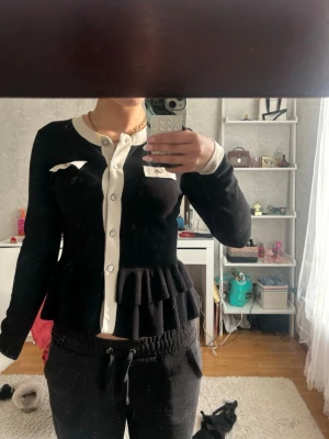 Svart peplumkofta från Seppälä Woman - Snygg svart kofta från Seppälä Woman med vita detaljer och knappar framtill. Koftan har peplumvolang i midjan, långa ärmar och kontrasterande vit kant vid hals, knappslå och fickor. Perfekt för dig som vill ha en chic och unik look.