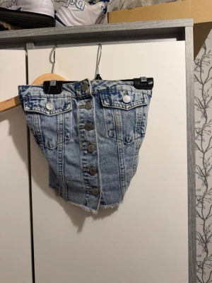Ljusblå denim tubtopp med knappar - Säljer en ljusblå tubtopp i denim med knappar framtill och rå kant nertill. Toppen har två bröstfickor med knappdetaljer och smockad rygg för extra bekväm passform. Perfekt för dig som vill ha en unik och edgy jeanslook.