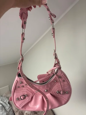 Rosa axelremsväska med nitar - Säljer en snygg rosa handväska i crocopräglat konstläder med silverfärgade nitar och detaljer. Väskan har en unik form, dragkedja upptill och en bred axelrem med metallspännen. Framtill finns dekorativa fickor och en matchande tag. Perfekt för dig som gillar statement-accessoarer.