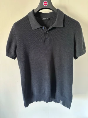 Mörkblå stickad pikétröja Sublevel - Mörkblå stickad pikétröja från Sublevel med korta ärmar och klassisk krage. Tröjan har tre knappar framtill och ribbade muddar vid ärmslut och nederkant. Perfekt för dig som gillar en enkel men stilren look.