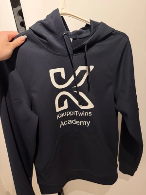 Mörkblå KauppiTwins Academy hoodie - Mörkblå hoodie med stor vit logga och texten KauppiTwins Academy på bröstet. Klassisk huva med dragsko och magficka framtill. Tillverkad i mjukt material som känns skönt mot huden. Perfekt för dig som gillar sportig och avslappnad stil. Jättebra skick nästan aldrig används