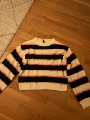 Randig stickad tröja i svart och vitt - Säljer en stickad tröja med breda svarta och vita ränder samt tunna rosa linjer. Tröjan har rund halsringning och långa ärmar. Perfekt för dig som gillar en avslappnad och trendig stil.