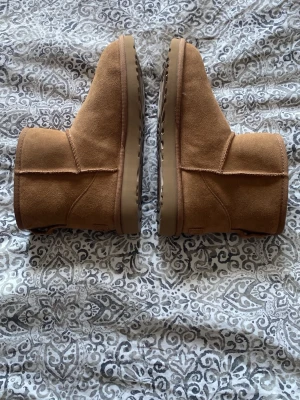 Klassiska bruna UGG-stövlar - Säljer ett par klassiska bruna UGG-stövlar i mocka med mjuk insida. De har en rund tå, mellanhögt skaft och grov sula för extra grepp. Perfekta för kalla dagar och riktigt sköna att ha på sig. Enkel och tidlös design som passar till det mesta.
