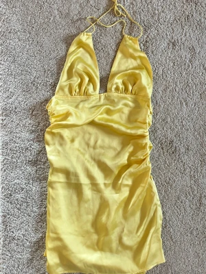 Gul halterneck klänning i satin - Snygg gul halterneck klänning i glansigt satinliknande material. Klänningen har smala knytband i nacken och djup urringning framtill. Kort modell med figurnära passform, perfekt för sommarens alla tillfällen. I fint skick!!
