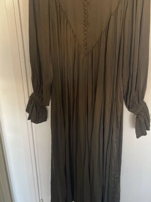 Långklänning med knappar och knyt - En långklänning i brun ton med knappar framtill och knyt vid ärmsluten. Klänningen har långärmade, lösa ärmar och ett plisserat fall. Perfekt för dig som gillar en bohemisk och avslappnad stil.