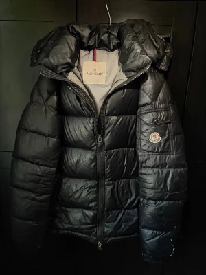 Svart dunjacka från Moncler - Snygg svart dunjacka från Moncler med klassisk puffer-design och huva. Jackan har Moncler-logga på ärmen, dragkedja framtill och praktiska fickor. Tillverkad i glansigt material och fodrad med dun för extra värme. Perfekt för kalla vinterdagar.
