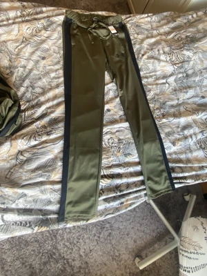 Olivgröna mjukisbyxor från Under Armour - Snygga olivgröna mjukisbyxor från Under Armour med svarta sidopaneler och dragsko i midjan. Tillverkade i ett mjukt och stretchigt material som ger skön passform. Perfekta för chill eller träning. Diskret logga på benet och smal passform. Passar folk som är 165-170