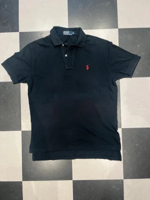 Svart pikétröja från Ralph Lauren - Klassisk svart pikétröja från Ralph Lauren med korta ärmar och krage. Tröjan har två knappar vid halsen och den ikoniska röda logotypen broderad på bröstet. Perfekt till jeans eller shorts för en stilren look.