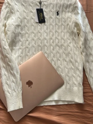 Vit kabelstickad tröja Polo Ralph Lauren - Kabelstickad vit tröja från Polo Ralph Lauren med klassisk rund hals och broderad logga på bröstet. Tröjan har långa ärmar och ribbade muddar vid ärmslut och nederkant. Perfekt för dig som gillar tidlös och stilren design.