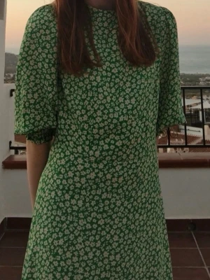 Grön blommig klänning med puffärm - Grön klänning med vitt blommönster och puffiga trekvartsärmar. Klänningen har en rak passform och rund halsringning. Perfekt för sommarens alla tillfällen och ger en fräsch och trendig look. Slitt i benet. 