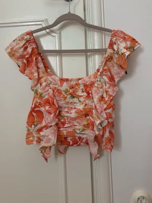 Blommig volangtopp i orange och vit - Supersöt blommig topp med volanger i orange och vitt. Toppen har breda axelband, smockad kropp och volangdetaljer framtill och baktill. Perfekt för sommardagar och har en luftig, croppad passform.