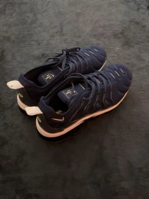Nike Air Max Plus blå sneakers - Snygga Nike Air Max Plus sneakers i mörkblå mesh och syntet med vita detaljer och unik svart sula med Air Max-dämpning. Ikonisk design med vågiga linjer och tydlig logga på sulan. Perfekta för dig som vill ha en sportig och trendig look.