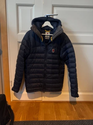 Mörkblå pufferjacka från Fjällräven - Snygg mörkblå pufferjacka från Fjällräven med huva och gul detalj vid dragkedjan. Jackan har en quiltad design, grå insida på huvan och klassisk Fjällräven-logga på bröstet. Perfekt för kalla dagar och riktigt skön att bära. Använd fåtal gånger och i väldigt fint skick, kvitto följer med. Hör av dig vid minsta fundering! Pris kan diskuteras 