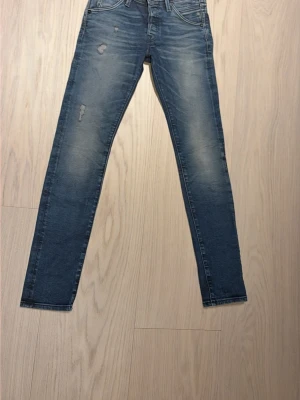 Blå slitna jeans från Jack & Jones - Säljer ett par blå jeans från Jack & Jones med skinny fit och slitningar på låret och knät. Jeansen har klassisk femficksdesign, knappgylf och snyggt tvättad look. Perfekta för en avslappnad och trendig stil.
