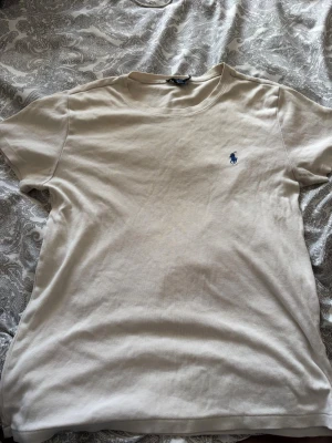 Vit t-shirt från Ralph Lauren - Snygg Vit t-shirt från Ralph Lauren med klassisk blå logga broderad på bröstet. Tillverkad i 100% bomull och har en enkel, stilren design med rund hals och korta ärmar. Perfekt för en clean och avslappnad look.