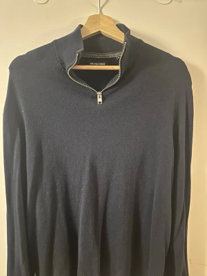 Mörkblå stickad tröja Jack & Jones - Mörkblå stickad tröja från Jack & Jones med halvzip och hög krage. Tröjan har en enkel och stilren design med långa ärmar och mjukt material som passar perfekt till lager på lager. Dragkedjan ger en sportig touch.