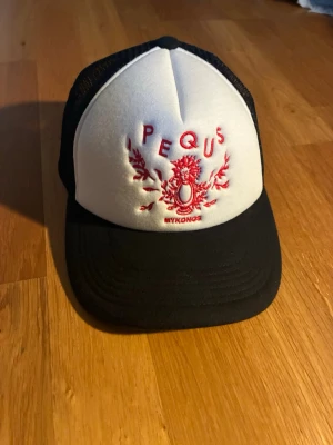 Svart och vit truckerkeps från PEQUS - Säljer en snygg svart och vit truckerkeps från PEQUS med röd brodyr framtill och texten 'MYKONOS'. Kepsen har mesh på sidorna och bak för extra ventilation samt böjd skärm. Perfekt för dig som vill ha en stilren och trendig accessoar.