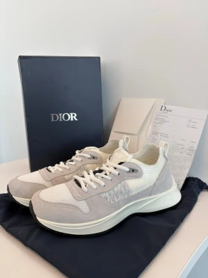Vita och grå Dior sneakers b25 - Snygga sneakers från Dior i vitt och ljusgrått med diskret logomönster på sidan. Skorna har en chunky sula i vitt och svart, meshdetaljer och mockaliknande partier. Perfekta för dig som vill ha en clean och exklusiv look.