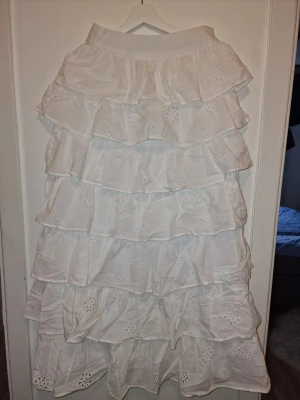 Vit volangkjol med anglaisebroderi - Viral kjol med flera lager volanger och fina broderade blommor. Den är gjord i ett lätt bomullstyg som känns luftigt och somrigt. Perfekt för dig som gillar romantisk och bohemisk stil. Supersnygg till studenten och midsommar. Passar även mig som har S i annat.