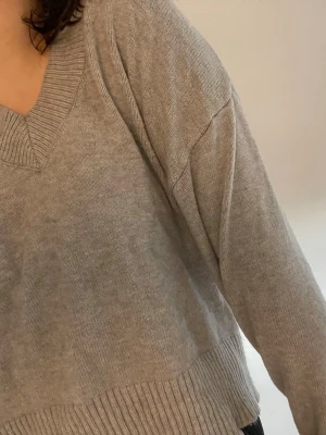 Grå stickad tröja med v-ringning - Mysig grå stickad tröja med bred ribbad mudd i nederkant och vid ärmslut. Tröjan har en djup v-ringning och avslappnad passform, perfekt för lager på lager. Materialet ser ut att vara mjukt och bekvämt, troligen bomull eller bomullsblandning.