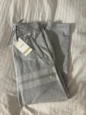 Grå mjukisbyxor från Stronger - Snygga ljusgrå mjukisbyxor från Stronger med två vita ränder och prickdetaljer på ena benet. Byxorna har dragsko i midjan och praktiska sidofickor. Dom är helt nya med prislapp 🌸