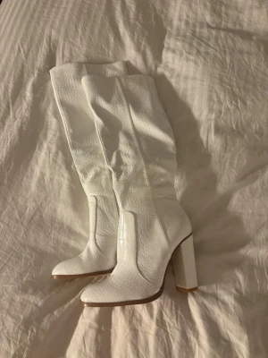 Vita höga boots med krokopräglat mönster - Säljer ett par helt oanvända vita höga stövlar med chunky klack och krokopräglat mönster. Stövlarna har rundad tå och dragkedja på insidan. Snygg retrovibe och passar perfekt till både kjol och jeans. Materialet är glansigt konstläder.
