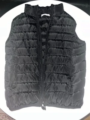 Svart dunväst med dragkedja - Snygg svart dunväst med horisontella sömmar och hög krage. Västen har dragkedja framtill och är tillverkad i ett glansigt material som ger en modern look. Perfekt att slänga över en hoodie eller tröja för extra värme. Priset kan diskuteras!!
