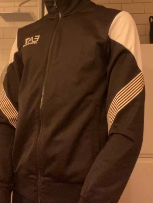 Svart och vit EA7 zip-jacka - Svart zip-jacka från EA7 Emporio Armani med vita paneler på axlar och armar samt vita ränder längs ärmarna. Jackan har krage, hel dragkedja och ribbade muddar. Perfekt för en sportig look. Även så säljs denna inte längre. Priset är inte fast hugget i sten. Skriv för frågor eller intresserad.