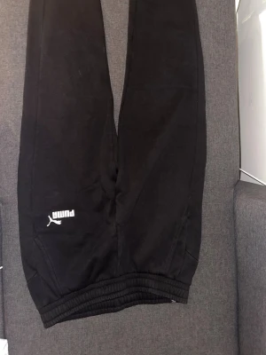 Svarta mjukisbyxor från Puma - Svarta mjukisbyxor från Puma med resår i midjan och benslut. Byxorna har en klassisk logga på vänster ben och är tillverkade i mjukt, fleeceliknande material. Perfekta för chill eller träning.