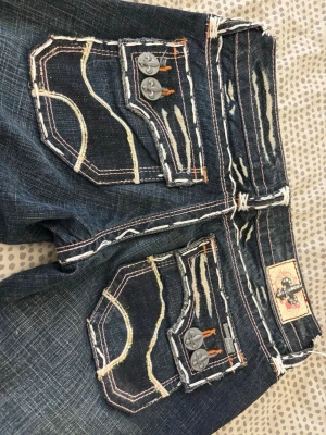Mörkblå Laguna Beach Jean Company utsvängda - Säljer ett par mörkblå jeans från Laguna Beach Jean Company med kraftiga kontrastsömmar, slitningar och dekorativa fickor med knappar. Jeansen har utsvängda ben och coola detaljer som broderier och patch bak. Perfekt för dig som gillar statement jeans! Dem är lite omsydda i utvängningen!💕💕