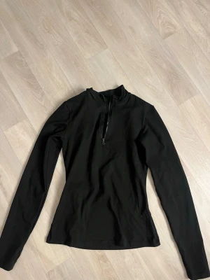 Svart långärmad tränings tröja med halv dragkedja - Stilren svart långärmad tröja med halv dragkedja framtill och hög krage. Tröjan har en figurnära passform och är tillverkad i ett mjukt, stretchigt material som passar perfekt till lager-på-lager. Enkel och clean design utan synligt märke.