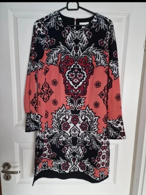 Mönstrad kort klänning - Säljer en kort klänning med långa ärmar och ett unikt blommigt mönster. Klänningen har rund halsringning och är tillverkad i ett mjukt, lätt material som känns skönt mot huden. 