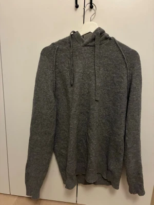 Grå stickad hoodie med huva från Replay - Mysig grå stickad hoodie med huva och dragsko från Replay, perfekt till våren. Tröjan har långa ärmar och ribbade muddar vid ärmslut och nederkant. Perfekt för dig som gillar en enkel och stilren look. Materialet känns mjukt mot huden och hoodien är i 55% Ull.