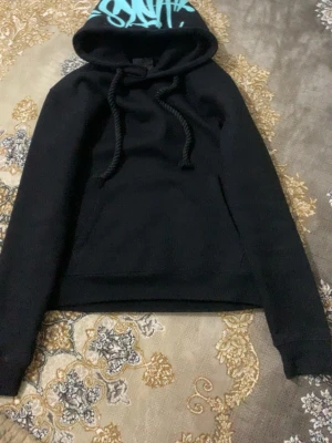 Svart hoodie med blå text på huvan - Säljer en svart hoodie med stor huva och breda snören. Huvan har ett blått tryck med text i graffiti-stil. Hoodien har en klassisk känguruficka framtill och ribbade muddar vid ärmslut och nederkant. Perfekt för en chill och avslappnad stil.