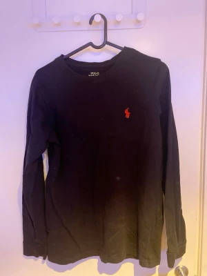 Svart långärmad tröja Polo Ralph Lauren - Klassisk svart långärmad tröja från Polo Ralph Lauren med rund halsringning och den ikoniska röda logotypen broderad på bröstet. Tröjan har en enkel och stilren design som passar till det mesta. Perfekt basplagg för garderoben.