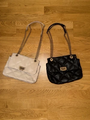 Svart och vit quiltad handväska med kedja - Två quiltade handväskor i svart och vitt med guldfärgade kedjeremmar och vridlås framtill. Klassisk rektangulär form och mjukt konstläder. Perfekt för dig som gillar stilrena accessoarer med trendig touch.