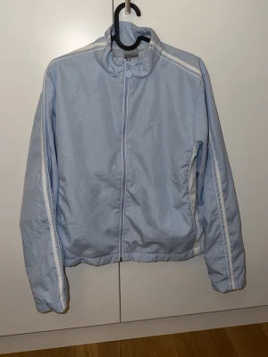 Ljusblå vindjacka från Nike - Ljusblå vindjacka från Nike med vita detaljer längs ärmar och sidor. Jackan har dragkedja framtill, hög krage och diskret Nike-logga på bröstet. Tillverkad i lätt och vindtätt material, perfekt för blåsiga dagar.